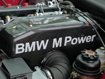 A detailed view of a BMW engine component featuring carbon fiber material with the text 'BMW M Power' prominently displayed. Various engine parts and components are visible around the main object, including cables, hoses, and connectors.