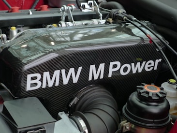 A detailed view of a BMW engine component featuring carbon fiber material with the text 'BMW M Power' prominently displayed. Various engine parts and components are visible around the main object, including cables, hoses, and connectors.