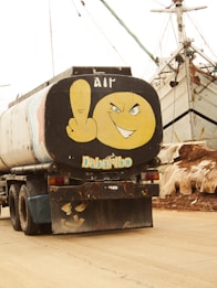 A truck with a tank displaying cartoon artwork on the back. The illustration features a large yellow face making a cheeky expression with one finger raised, situated against the backdrop of boats and industrial equipment.