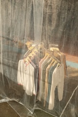 A collection of shirts and tops on hangers is visible through a translucent plastic curtain. The clothes are arranged neatly on a rack and vary in colors, including white, blue, and beige. The background appears to be an industrial or weathered wall, adding a rustic feel.