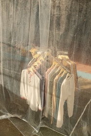 A collection of shirts and tops on hangers is visible through a translucent plastic curtain. The clothes are arranged neatly on a rack and vary in colors, including white, blue, and beige. The background appears to be an industrial or weathered wall, adding a rustic feel.