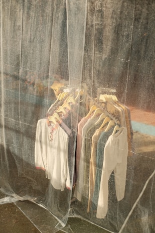 A collection of shirts and tops on hangers is visible through a translucent plastic curtain. The clothes are arranged neatly on a rack and vary in colors, including white, blue, and beige. The background appears to be an industrial or weathered wall, adding a rustic feel.