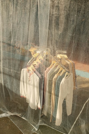 A collection of shirts and tops on hangers is visible through a translucent plastic curtain. The clothes are arranged neatly on a rack and vary in colors, including white, blue, and beige. The background appears to be an industrial or weathered wall, adding a rustic feel.