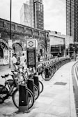 A row of rental bicycles is lined up on a street sidewalk. In the background, there are tall modern buildings and an old brick wall covered with graffiti. People are interacting near the bicycles, and a sign with a transport logo is visible.
