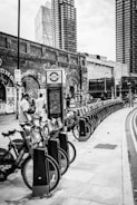 A row of rental bicycles is lined up on a street sidewalk. In the background, there are tall modern buildings and an old brick wall covered with graffiti. People are interacting near the bicycles, and a sign with a transport logo is visible.
