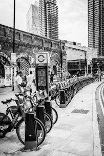 A row of rental bicycles is lined up on a street sidewalk. In the background, there are tall modern buildings and an old brick wall covered with graffiti. People are interacting near the bicycles, and a sign with a transport logo is visible.