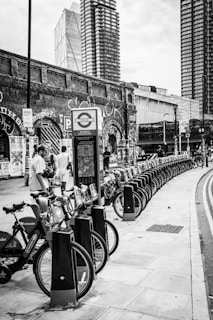 A row of rental bicycles is lined up on a street sidewalk. In the background, there are tall modern buildings and an old brick wall covered with graffiti. People are interacting near the bicycles, and a sign with a transport logo is visible.