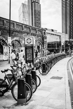 A row of rental bicycles is lined up on a street sidewalk. In the background, there are tall modern buildings and an old brick wall covered with graffiti. People are interacting near the bicycles, and a sign with a transport logo is visible.