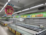 A nearly empty supermarket with empty shelves and freezer units. Colorful signs advertise discounts of up to 80% and other promotional offers in bold fonts above the shelves. The image conveys a sense of clearance sale or store closure.