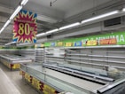 A nearly empty supermarket with empty shelves and freezer units. Colorful signs advertise discounts of up to 80% and other promotional offers in bold fonts above the shelves. The image conveys a sense of clearance sale or store closure.
