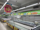 A nearly empty supermarket with empty shelves and freezer units. Colorful signs advertise discounts of up to 80% and other promotional offers in bold fonts above the shelves. The image conveys a sense of clearance sale or store closure.
