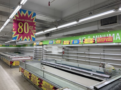 A nearly empty supermarket with empty shelves and freezer units. Colorful signs advertise discounts of up to 80% and other promotional offers in bold fonts above the shelves. The image conveys a sense of clearance sale or store closure.