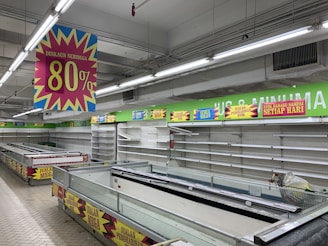 A nearly empty supermarket with empty shelves and freezer units. Colorful signs advertise discounts of up to 80% and other promotional offers in bold fonts above the shelves. The image conveys a sense of clearance sale or store closure.