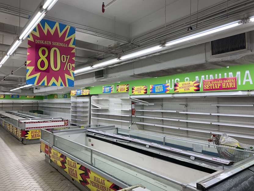 A nearly empty supermarket with empty shelves and freezer units. Colorful signs advertise discounts of up to 80% and other promotional offers in bold fonts above the shelves. The image conveys a sense of clearance sale or store closure.