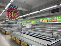 A nearly empty supermarket with empty shelves and freezer units. Colorful signs advertise discounts of up to 80% and other promotional offers in bold fonts above the shelves. The image conveys a sense of clearance sale or store closure.