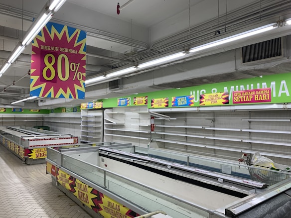 A nearly empty supermarket with empty shelves and freezer units. Colorful signs advertise discounts of up to 80% and other promotional offers in bold fonts above the shelves. The image conveys a sense of clearance sale or store closure.