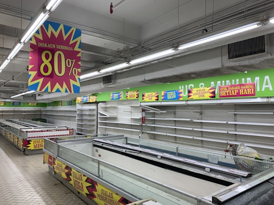 A nearly empty supermarket with empty shelves and freezer units. Colorful signs advertise discounts of up to 80% and other promotional offers in bold fonts above the shelves. The image conveys a sense of clearance sale or store closure.