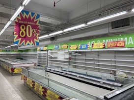 A nearly empty supermarket with empty shelves and freezer units. Colorful signs advertise discounts of up to 80% and other promotional offers in bold fonts above the shelves. The image conveys a sense of clearance sale or store closure.