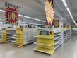 Empty supermarket shelves are surrounded by bright sale signs displaying discounts up to 60%. The banners are in bold colors, with phrases indicating a significant stock clearance. The store appears vacant, with no products on the shelves and no people present.