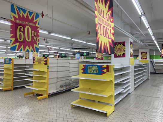 Empty supermarket shelves are surrounded by bright sale signs displaying discounts up to 60%. The banners are in bold colors, with phrases indicating a significant stock clearance. The store appears vacant, with no products on the shelves and no people present.