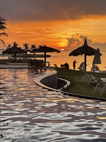 A tranquil scene featuring a sunset over a calm ocean. Silhouettes of palm trees and thatched umbrellas line the shore, adding to the picturesque setting. The water reflects the vibrant hues of the sunset, creating a serene and peaceful atmosphere. A couple of lounge chairs are positioned on the grassy area, overlooking the view.