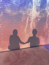 Two silhouettes standing side by side on a background featuring a blend of pink and blue hues. They are holding hands, casting shadows against a colorfully lit wall, creating a romantic and abstract visual effect.