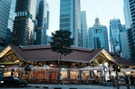 A vibrant cityscape with a hawker center in the foreground surrounded by a cluster of modern skyscrapers. The dining area features lights draped across the outdoor seating, enhancing the lively atmosphere. The buildings in the background display a mix of architectural styles and heights.