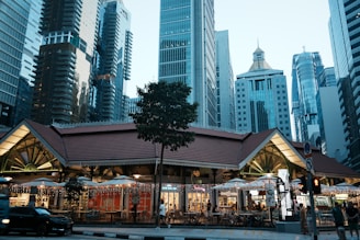 A vibrant cityscape with a hawker center in the foreground surrounded by a cluster of modern skyscrapers. The dining area features lights draped across the outdoor seating, enhancing the lively atmosphere. The buildings in the background display a mix of architectural styles and heights.