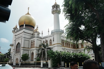 A grand mosque features a large golden dome and a tall minaret. The building is surrounded by lush green trees and ornate architectural details. Several small plants are visible near the entrance, and a few people can be seen near the bottom of the image.