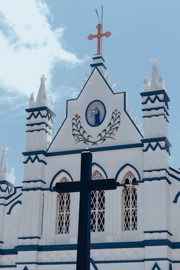 A white church facade with pointed architectural elements is adorned with blue accents and a large black cross in the foreground. A depiction of a religious figure is set within a circular frame, decorated with a floral motif above the entrance. The sky is bright and partly cloudy.