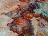 Aerial view of a striking geological formation with swirling, concentric patterns in vibrant shades of rust, brown, and turquoise. The textures are rich and layered, creating a natural artwork reminiscent of a topographical map.