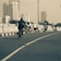 A group of cyclists ride along a city road, with tall buildings visible in the background. The sky is overcast, and the scene appears to have a sepia or vintage filter applied. The road is divided by white lines and bordered by a stone railing.