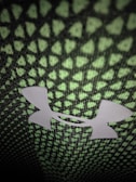 A close-up of a textured fabric with a repeating pattern of small green triangles on a black background. In the center, there is a prominent white logo of intersecting shapes.