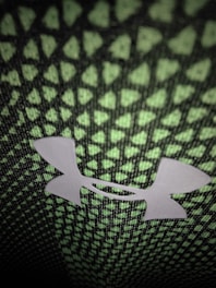 A close-up of a textured fabric with a repeating pattern of small green triangles on a black background. In the center, there is a prominent white logo of intersecting shapes.