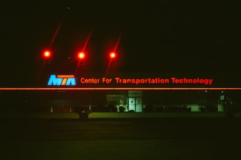 A building exterior features a large illuminated sign reading 'Center For Transportation Technology' in red neon letters with an 'MTA' logo in blue, white, and red on the left side. Above the sign, three red lights cast a glow against the dark night sky. The building appears to have reflective glass windows and a centrally positioned entrance with closed doors.