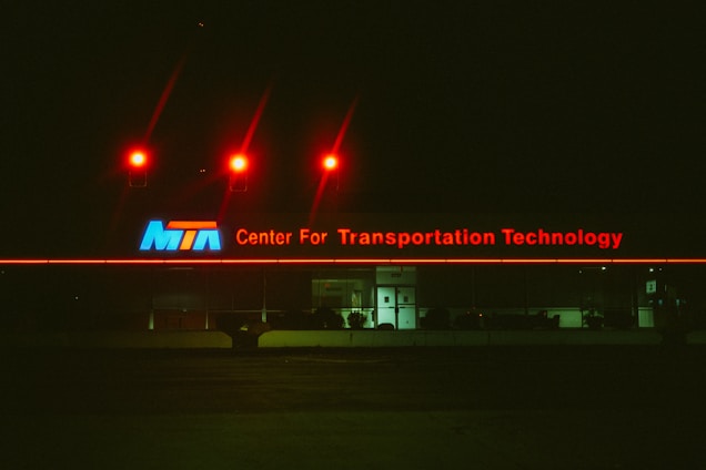 A building exterior features a large illuminated sign reading 'Center For Transportation Technology' in red neon letters with an 'MTA' logo in blue, white, and red on the left side. Above the sign, three red lights cast a glow against the dark night sky. The building appears to have reflective glass windows and a centrally positioned entrance with closed doors.