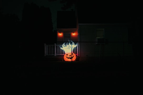A nighttime scene featuring a small suburban house with a white railing and a porch. Orange lights illuminate the front door area, casting a warm glow. In the foreground, a bright, illuminated Halloween decoration with a ghost and a jack-o'-lantern is prominently displayed.