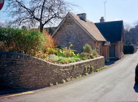A quaint stone cottage is surrounded by a lush garden filled with colorful flowers and greenery. The stone wall lines the property, and a tree stands bare against the blue sky. The road adjacent to the cottage suggests a quiet, rural setting.