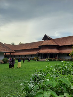 A traditional building with a large sloping tiled roof is seen on overcast day. The structure is surrounded by a well-maintained lawn and lush green plants. Several people are gathered on the lawn, some in groups, indicating a social or casual gathering.
