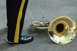 A shiny brass French horn is lying on a paved surface next to a person wearing black pants with a yellow stripe and polished black shoes.