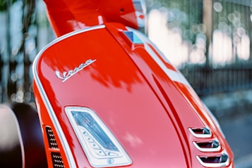 A close-up view of a red Vespa scooter showcasing its glossy finish and emblem. The background is softly blurred, emphasizing the sleek design and bright color of the scooter.