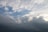 Cloudy sky featuring a mix of cumulus and stratus clouds with patches of blue sky peeking through. The light from the sun subtly illuminates the edges of the clouds, creating a dynamic contrast between the bright and shadowed areas.