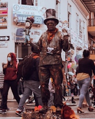 A street performer dressed in a painted suit and wearing a top hat stands on a platform. He is holding two puppets, one in each hand, and has a camera hanging around his neck. The background is a busy street with people walking by, some wearing masks. The scene is lively with various signs and storefronts visible.