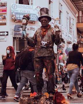 A street performer dressed in a painted suit and wearing a top hat stands on a platform. He is holding two puppets, one in each hand, and has a camera hanging around his neck. The background is a busy street with people walking by, some wearing masks. The scene is lively with various signs and storefronts visible.