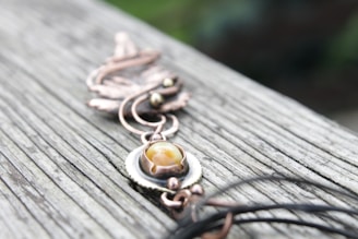 A detailed close-up of a piece of jewelry designed with copper-colored metal and a central yellow stone, placed on a weathered wooden surface. The design appears intricate with swirls and a leaf-like pattern.
