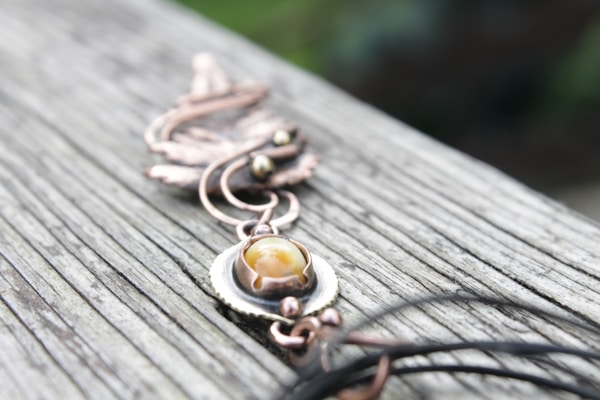 A detailed close-up of a piece of jewelry designed with copper-colored metal and a central yellow stone, placed on a weathered wooden surface. The design appears intricate with swirls and a leaf-like pattern.