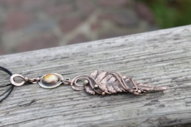 A handcrafted necklace with an intricate design lies on a weathered wooden surface. The necklace features a metal pendant with leaf motifs and a polished stone centerpiece. The metal has a warm, coppery tone with a delicate chain attached.