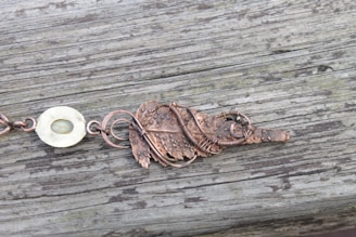 An intricately designed metallic leaf-shaped ornament with a rustic appearance rests on a wooden surface with visible grain. The metal features textured details and a spiral wire wrapping around it. To the left, a rounded, pale stone-like element is connected to the metal piece.