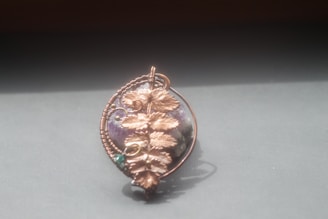 A handcrafted pendant featuring a large stone encased in intricate copper wirework that forms a design resembling leaves. The pendant is prominently displayed against a plain background and appears to be lit by natural light.