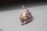 An intricately designed metal pendant with a swirling pattern and leaf motif, set against a muted background. The pendant incorporates a blend of copper wire and a brass-colored leaf shape, along with a textured purple stone.